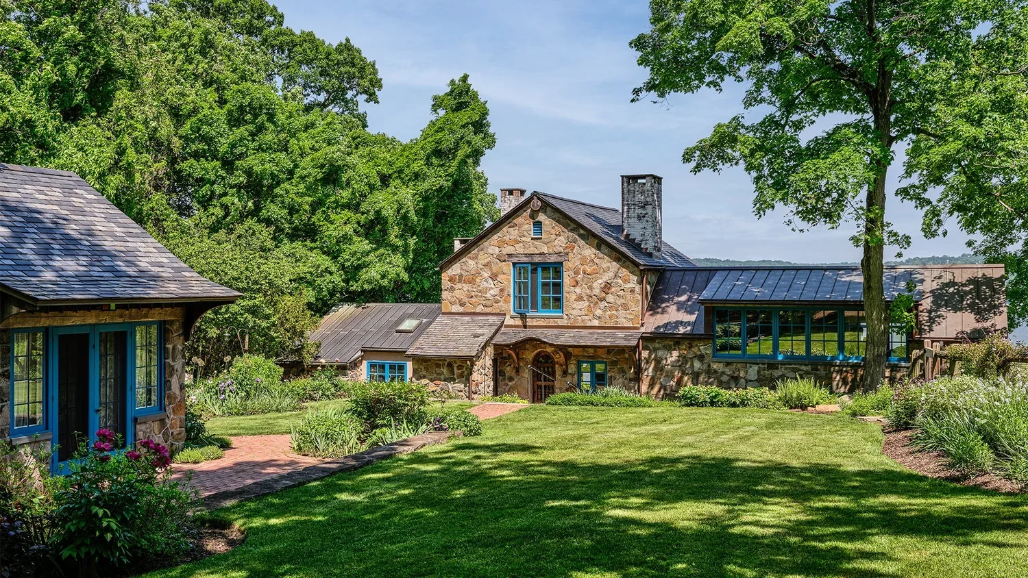 A scenic view of a large stone house with a metal roof, features blue window frames, surrounded by lush green trees and a well-maintained lawn.