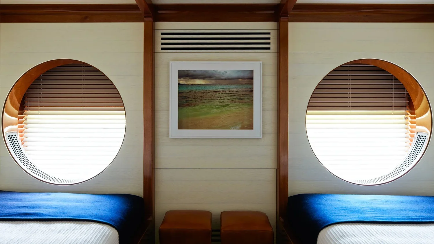 Interior of a room with two beds with blue covers, round windows with wooden frames, a framed beach scene painting on the wall, and small wooden benches between the beds.