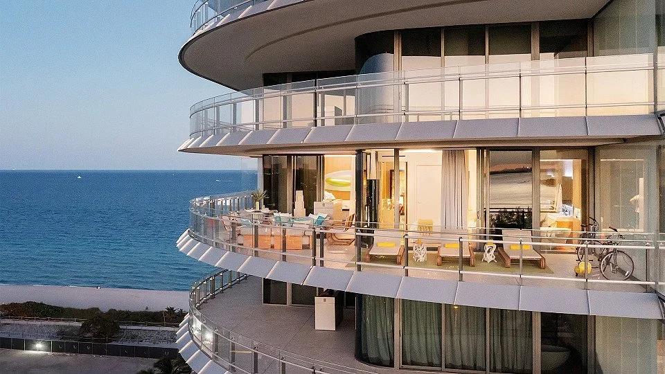 Luxury apartment balcony overlooking the ocean, with patio furniture and bicycle, during sunset.