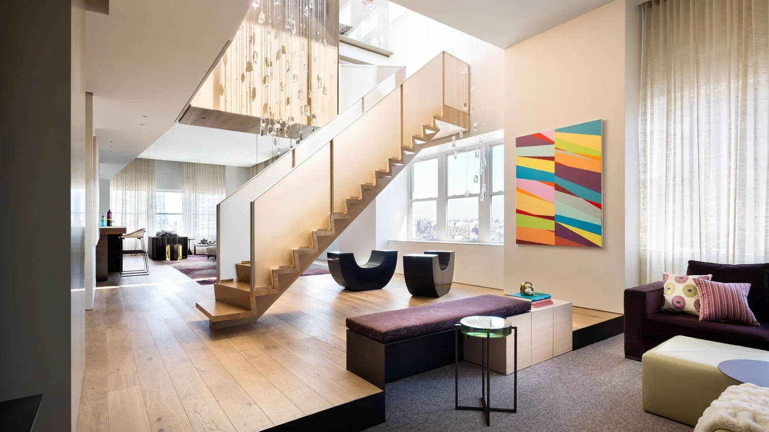 Modern living room with a staircase, colorful wall art, large windows with sheer curtains, and contemporary furniture including a purple sofa with patterned pillows, a bench, and a small side table.