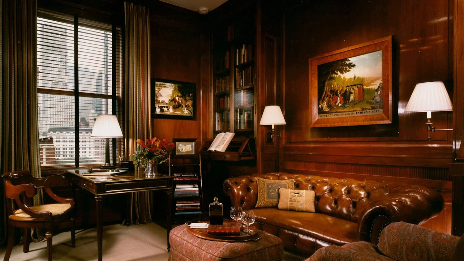 A cozy traditional living room with dark wood paneling, a leather chesterfield sofa, a window with blinds and striped curtains, a small round table with glasses and a tray, a bouquet of flowers, two table lamps, and framed artwork on the walls.
