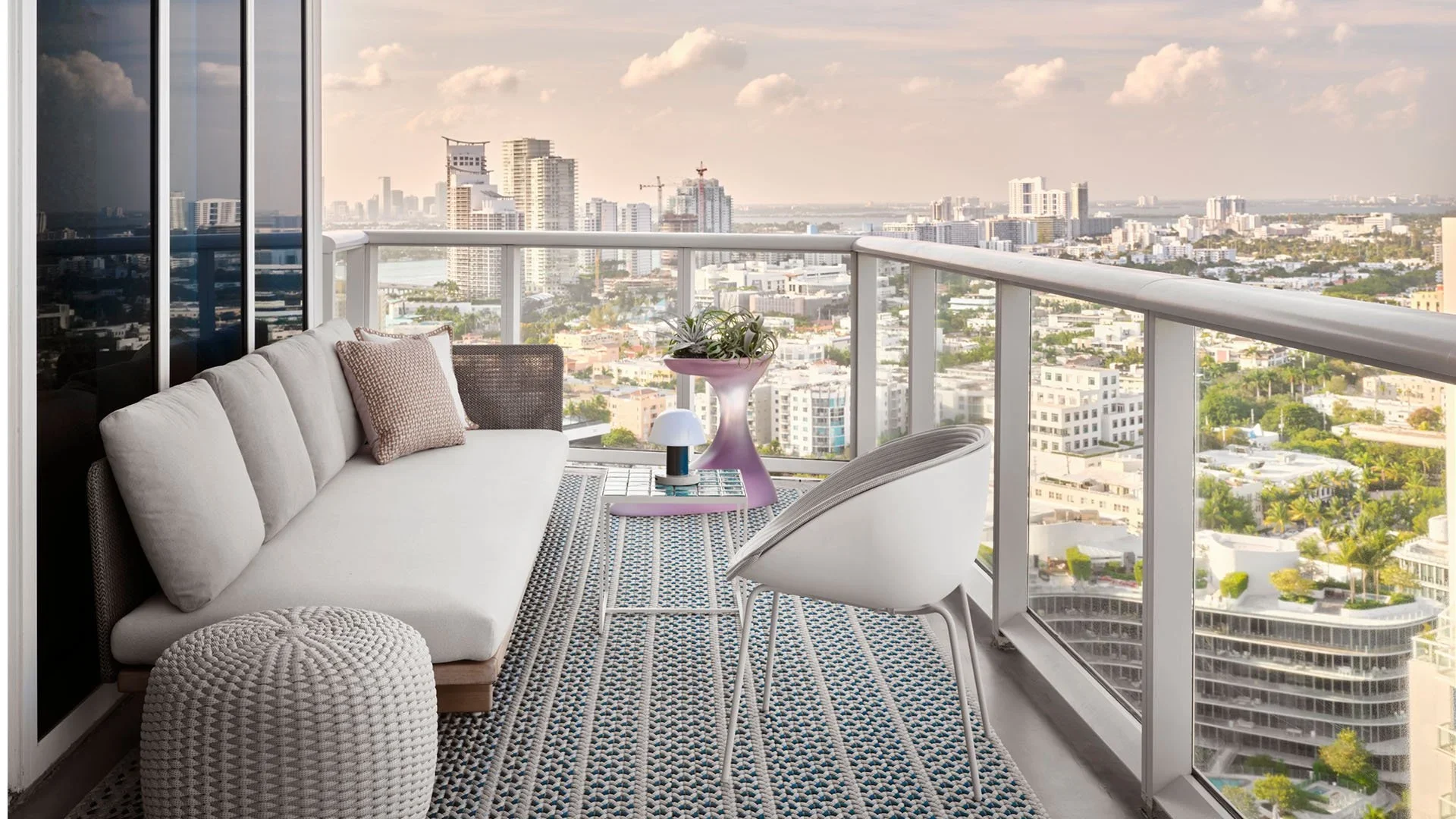 Balcony with white sofa, beige pillows, knitted pouf, white and gray chair, purple side table with green plants, and city skyline view with tall buildings and cloudy sky.