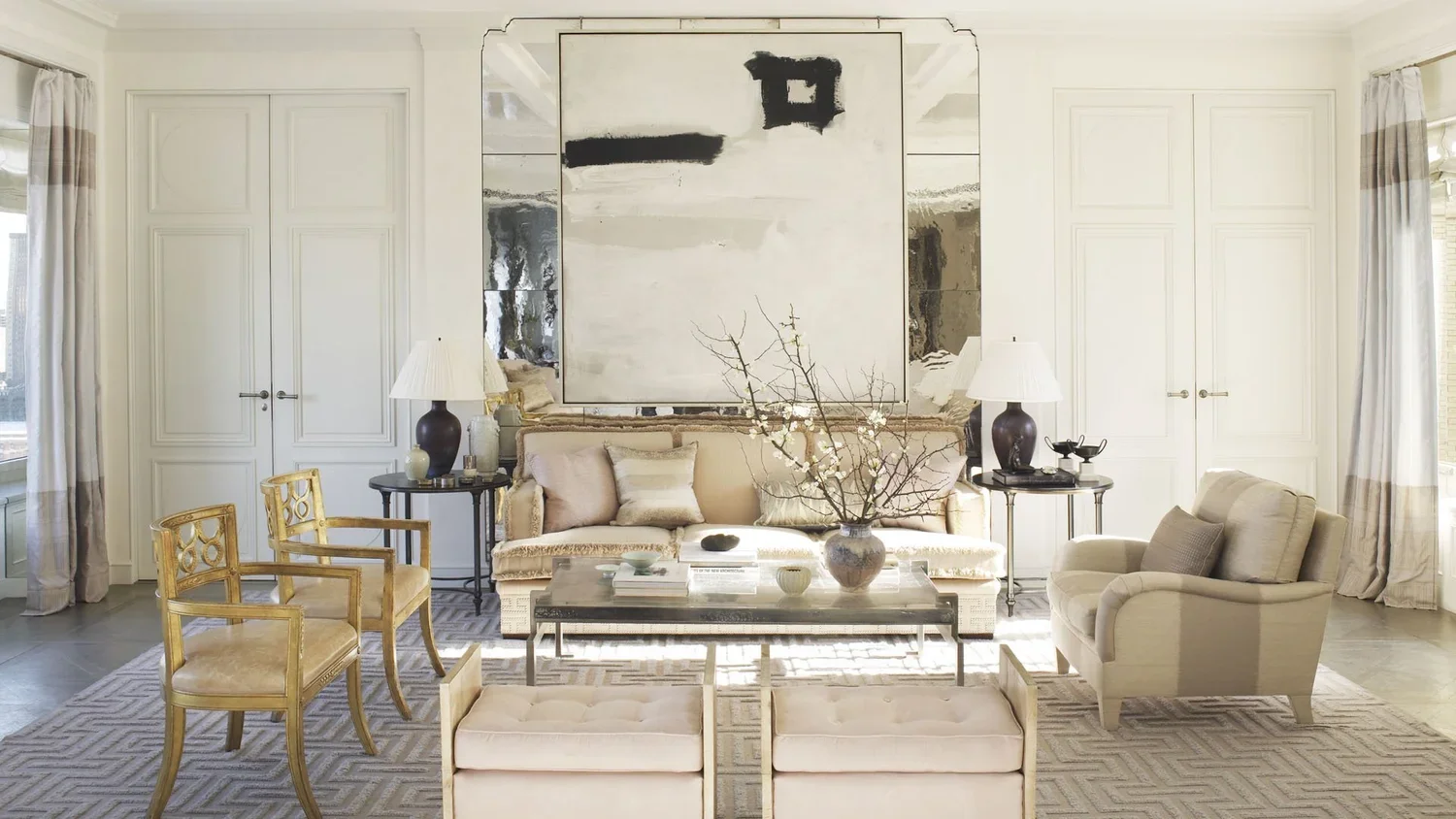 Living room with a cream-colored sofa, a glass coffee table with books and a vase with branches, matching armchair, side tables with lamps, gold chairs, and large abstract artwork on the wall. Natural light from the windows illuminates the space.