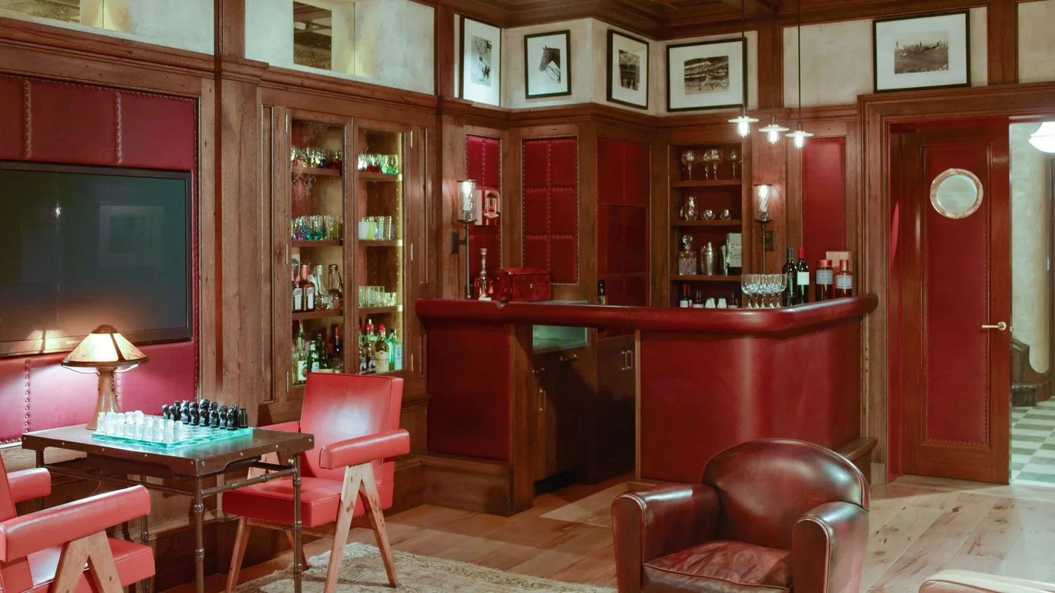 A cozy home bar with wooden paneling, glass shelves with liquor bottles, red leather chairs, a chess set on a table, wine glasses, and framed vintage photos on the wall.