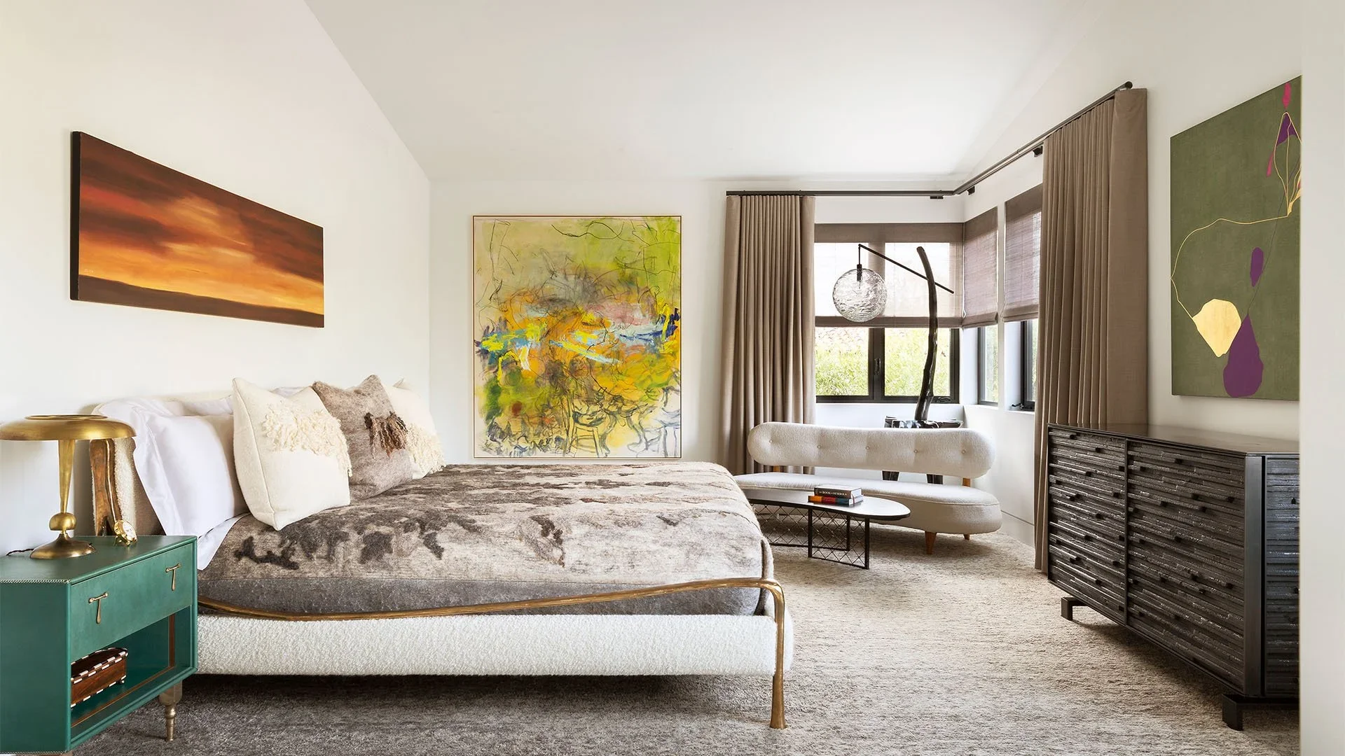 A well-decorated bedroom with a bed, artwork, a sofa, a black sideboard, and large windows with beige curtains