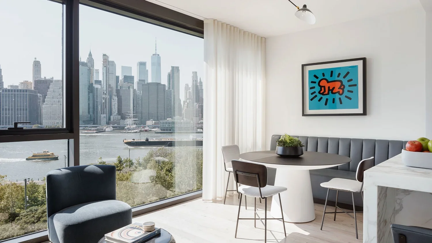 Modern apartment living room with large window overlooking Manhattan skyline, featuring a round black and white dining table with chairs, gray bench, and abstract art on white wall.