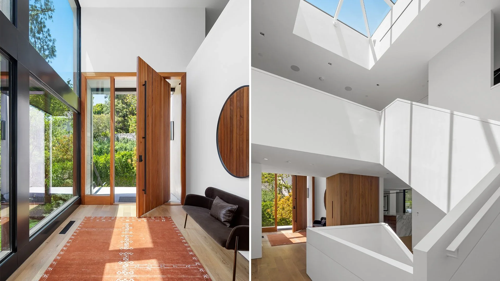 Modern interior with large floor-to-ceiling glass windows, wooden front door, black sofa, and orange rug on light wood flooring, with greenery visible outside.