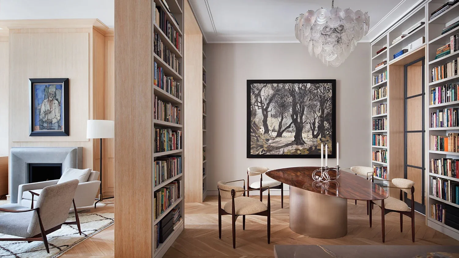 A modern living room with a round wooden table and six chairs, framed artwork on the wall, bookshelves filled with books, a chandelier, and a sitting area with a fireplace and armchair.