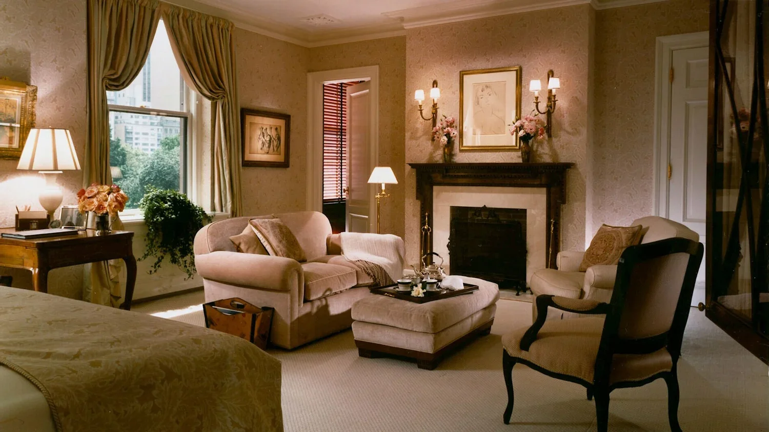 A cozy living room with a fireplace, a beige sofa, an armchair, a coffee table with tea set, a window with curtains, framed artwork, and decorative lamps.