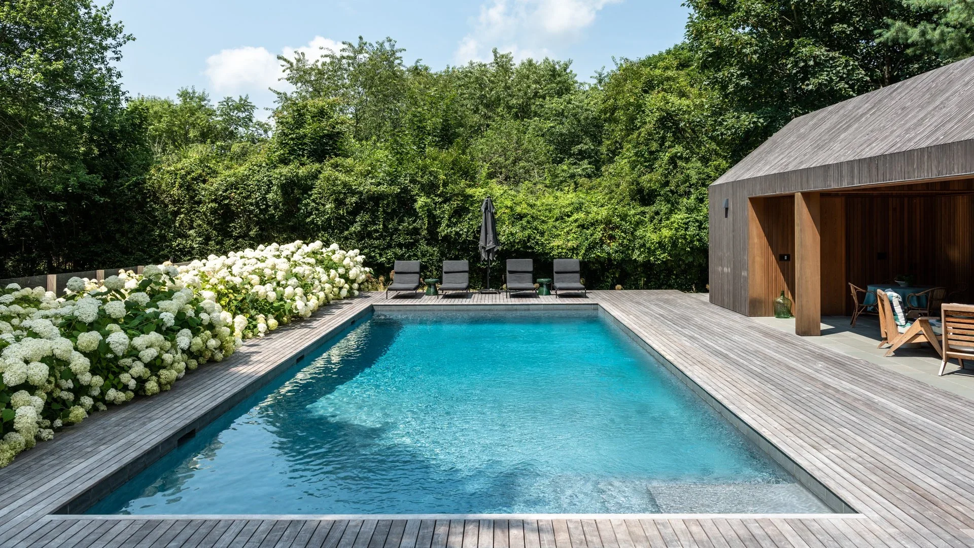 Outdoor swimming pool with clear blue water, surrounded by a wooden deck, white hydrangea bushes, four black lounge chairs with an umbrella, and a modern wooden structure with seating area, set against lush green trees on a sunny day.