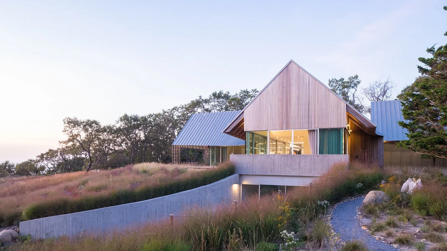 Modern house with large glass windows and a sloped metal roof, situated on a hillside with natural landscaping and a winding gravel path.