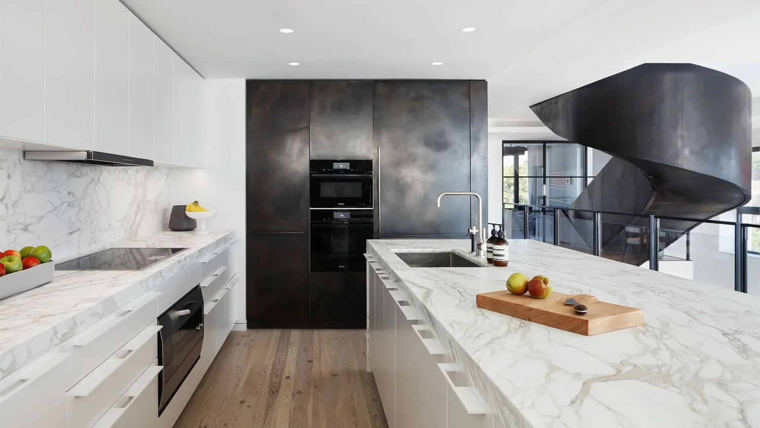 Modern kitchen with white marble countertops, black appliances, and a unique black spiral staircase in the background.