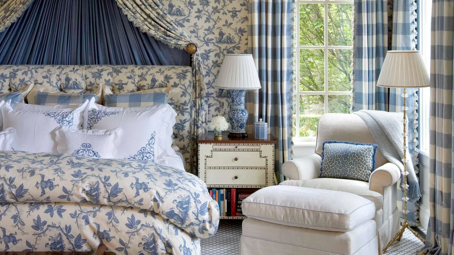 A bedroom with blue and white floral and plaid decor, featuring a large bed with a tufted headboard, several white pillows, a small white armchair with a, cushion, two blue lamps with white shades on bedside tables, a window with blue and white plaid