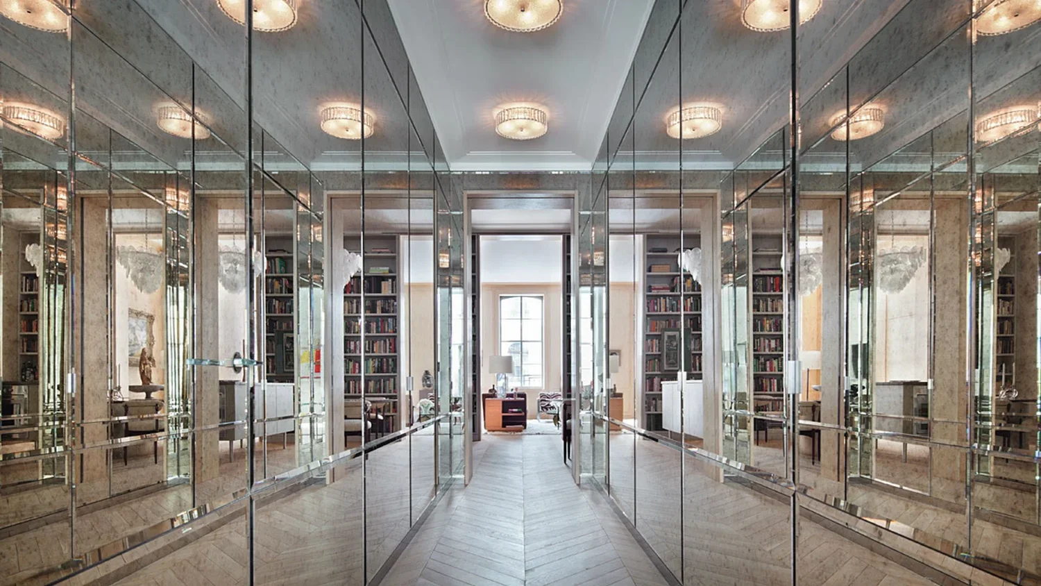 Mirror-lined hallway leading to a room with bookshelves and a large window at the end, with ceiling lights reflecting in the mirrors.