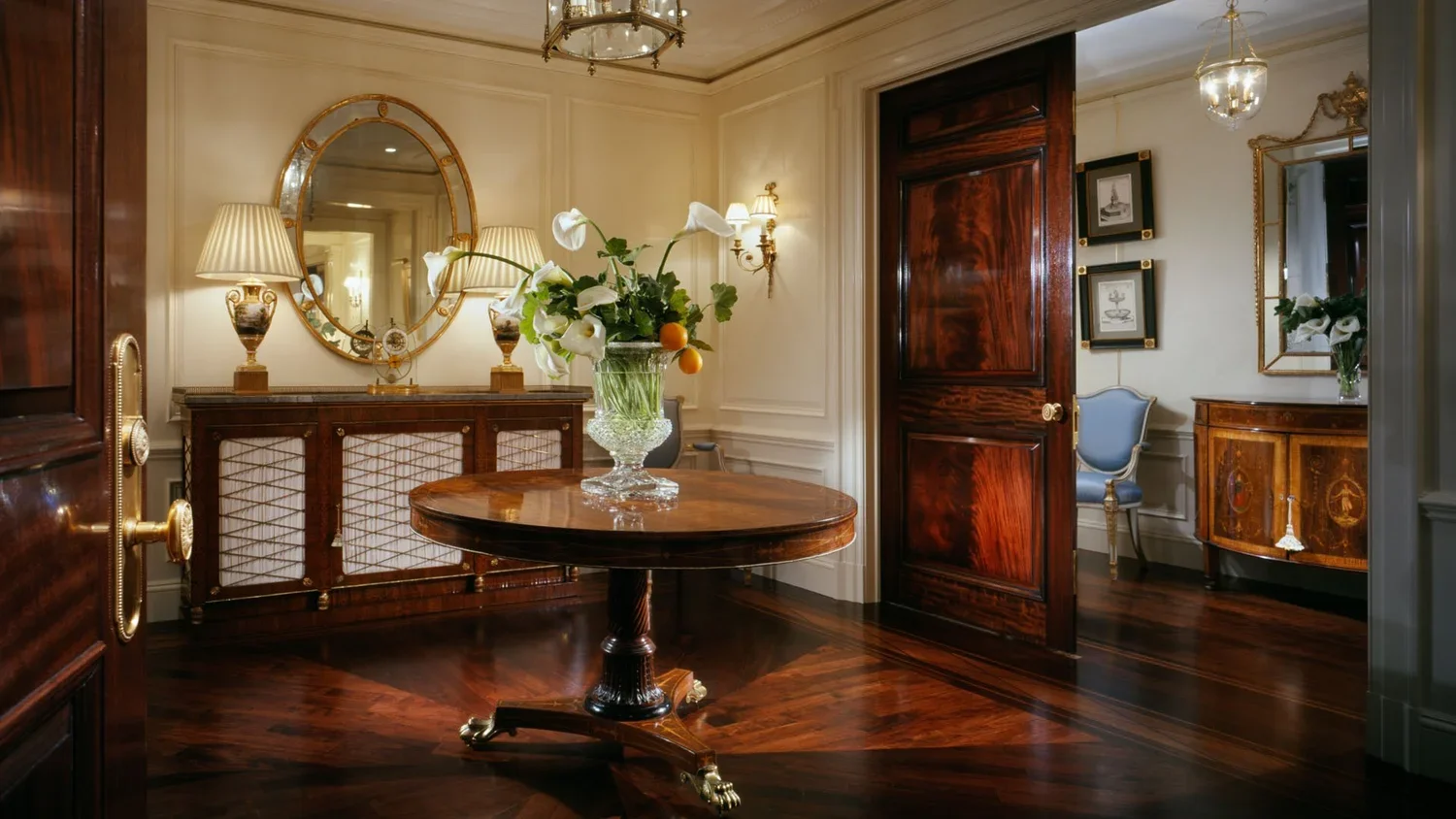 Elegant antique dining room with dark wood furniture, including a round table with a floral arrangement, a sideboard, and a small cabinet. The room features cream walls, decorative lighting, and a wood floor with a geometric pattern. A doorway leads 