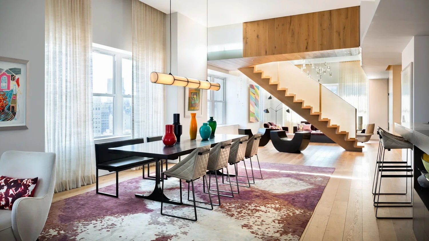 Modern living and dining area with large windows, curtains, colorful vases on table, wooden staircase, artwork on walls, and contemporary furniture.