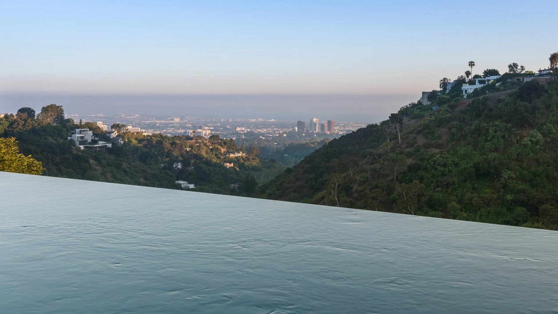 A view of a valley with green hills and residential houses. In the background, there is a cityscape with tall buildings and a clear blue sky.