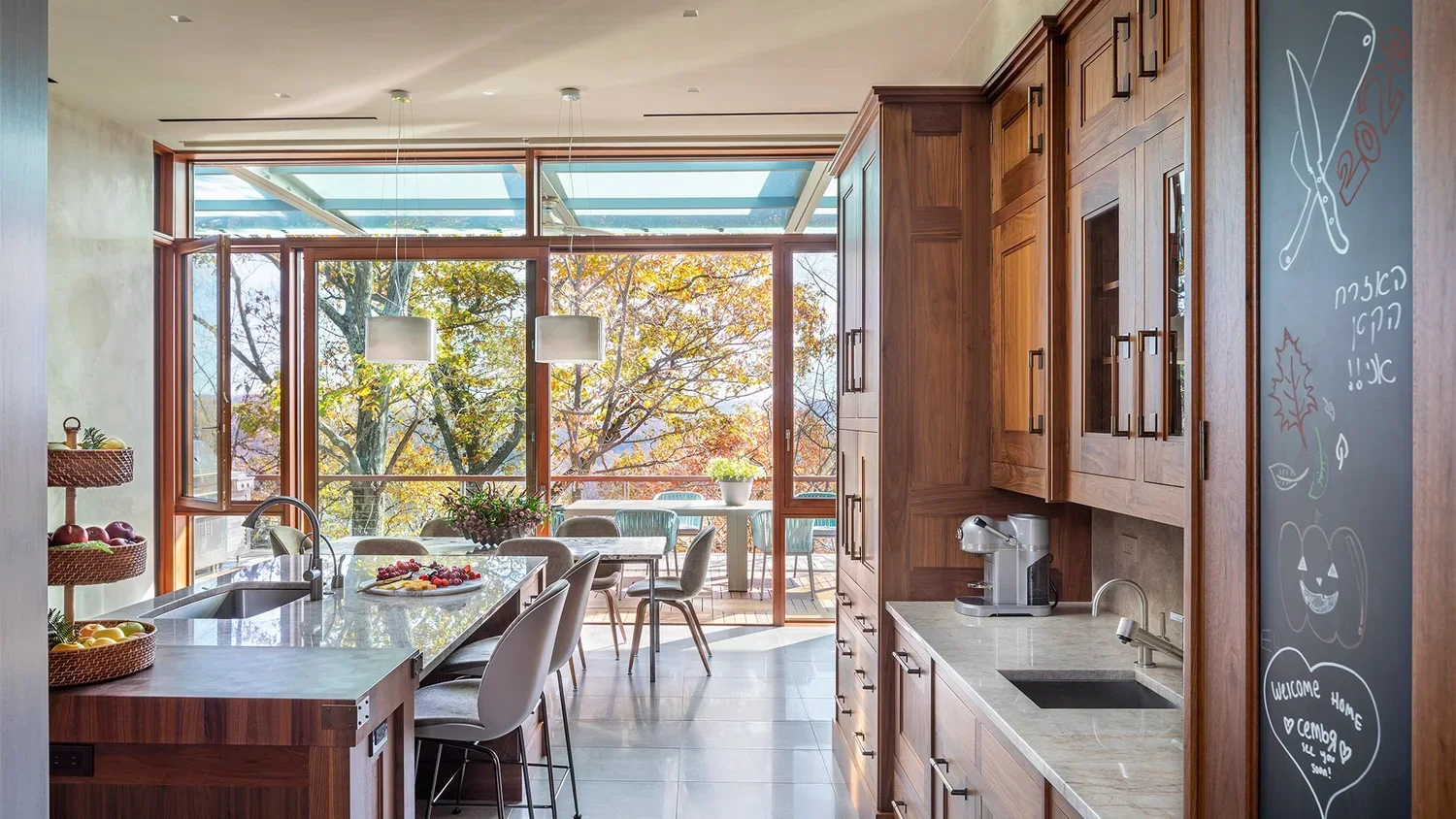 Modern kitchen with large windows, wooden cabinets, a kitchen island, and a view of an outdoor patio with trees in fall.