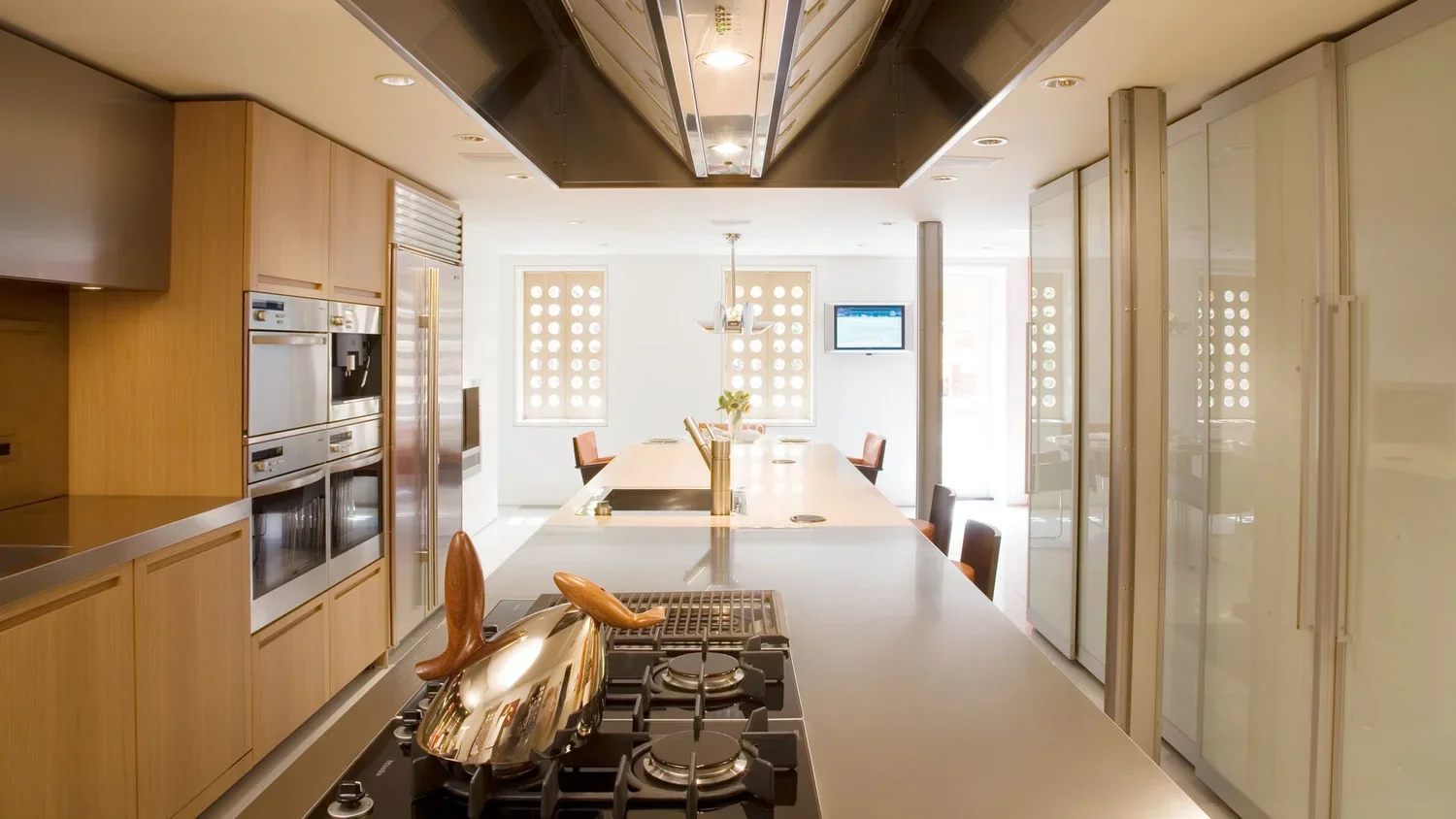 Modern kitchen with wooden cabinets, stainless steel appliances, a large central island with a stovetop, and chairs around the island. Bright sunlight streaming through windows.