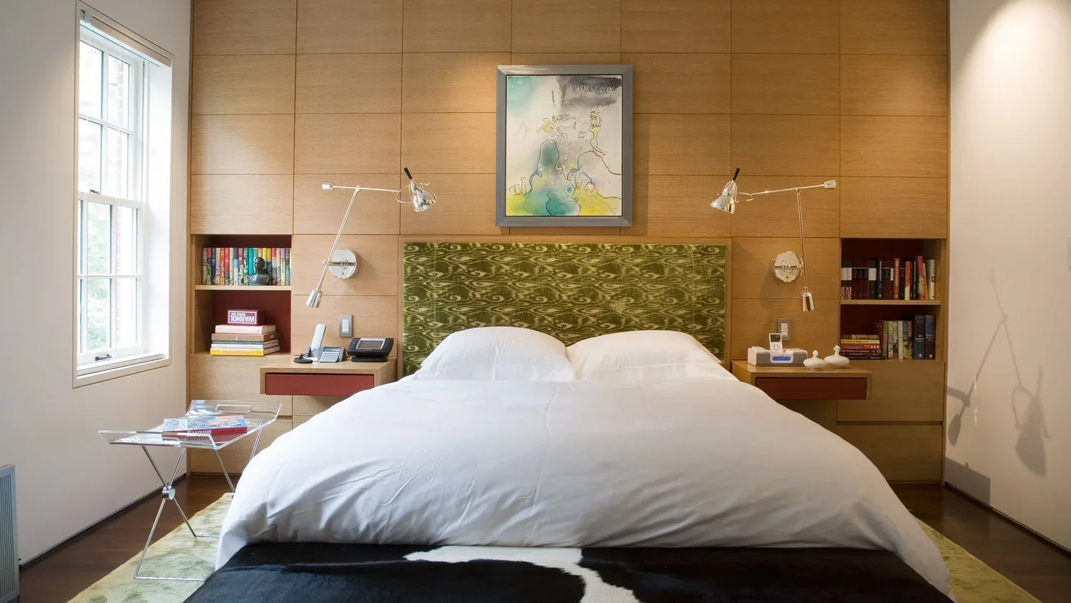 A modern bedroom with a bed, two bookshelves on each side of the headboard, and wall-mounted lamps. There are two wall-mounted lamps above the headboard, a window to the left, and a piece of abstract art hanging above the headboard. A transparent sid