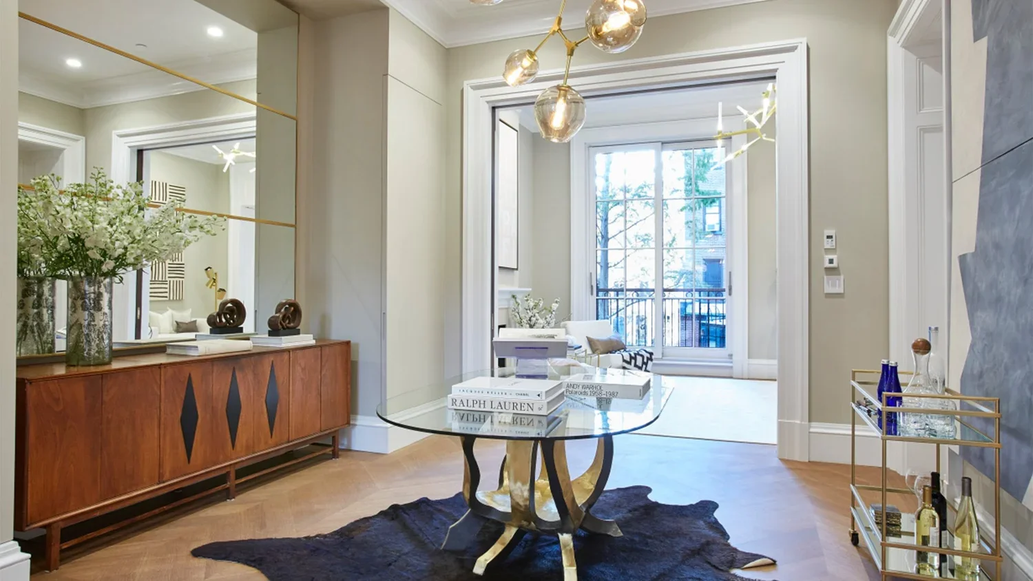 Living room with glass table on a black cowhide rug, wooden sideboard with decorative objects and flowers, gold and glass bar cart, and large window with balcony view.