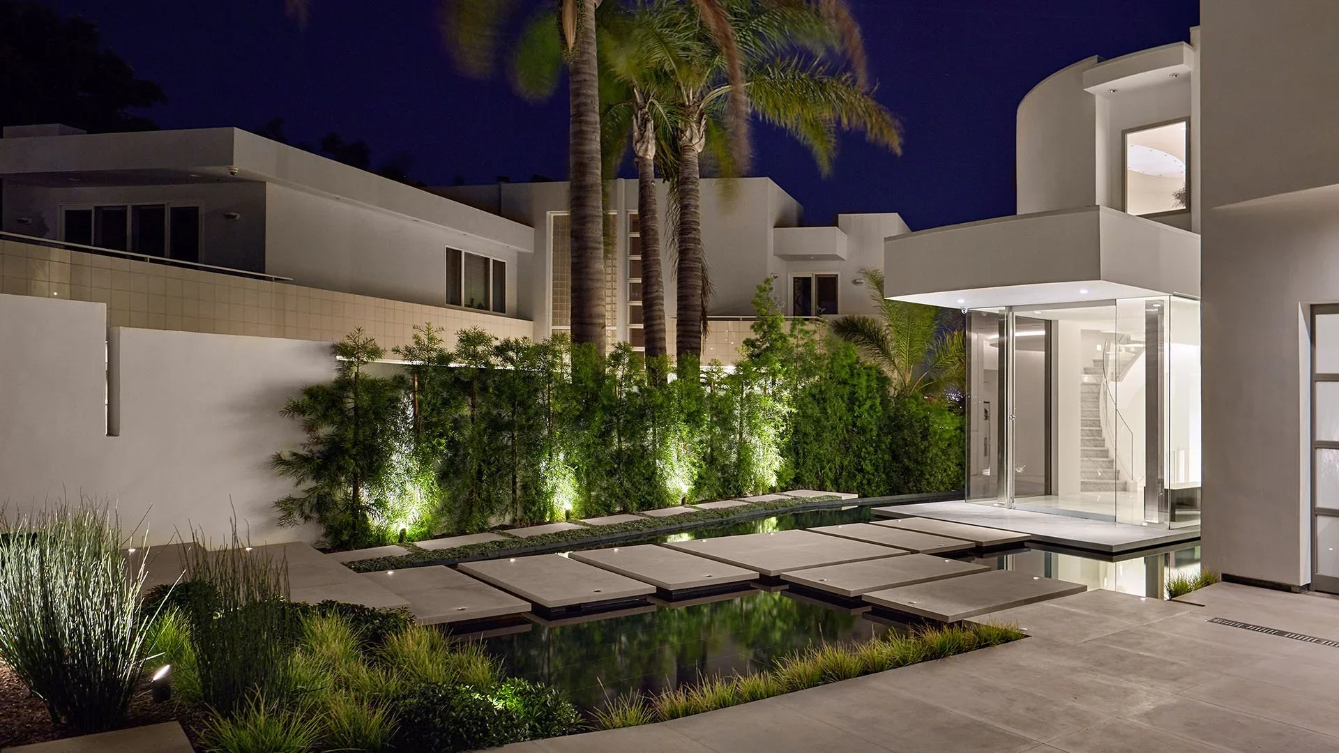 Modern house exterior at night with illuminated plants, stepping stones over a reflecting pond, and a glass entrance door.
