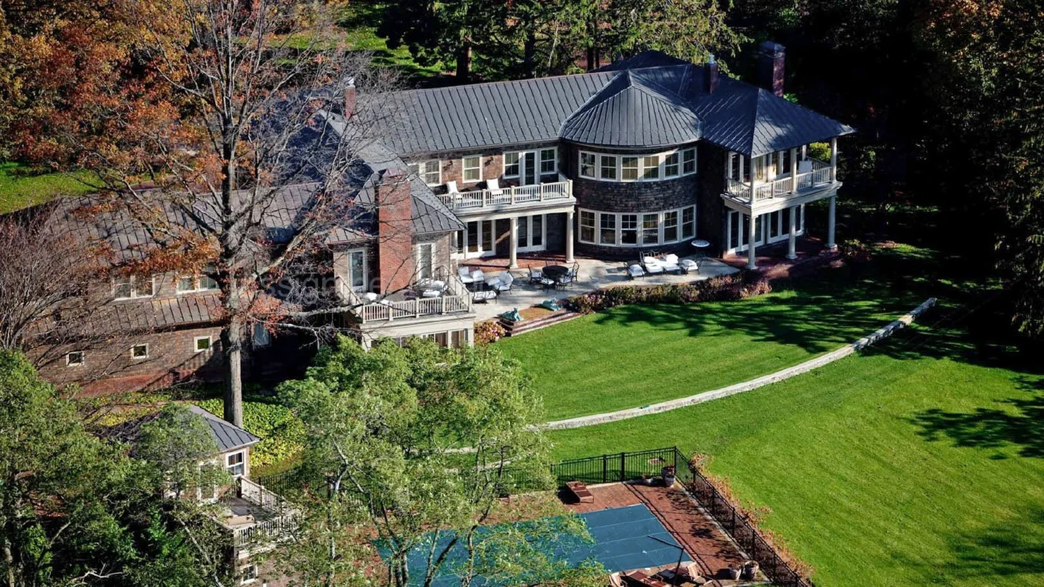 A large, multi-story house with a gray metal roof, brick and stone exterior, and multiple balconies, situated in a lush, green backyard with a well-maintained lawn, trees, and outdoor furniture.