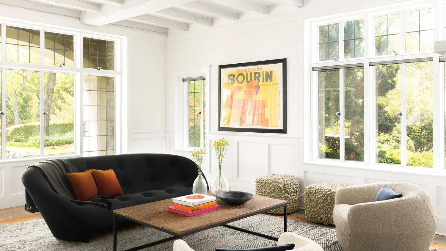 Bright living room with large windows, white walls, and modern furniture including a black sofa with orange pillows, a round beige armchair, a rustic coffee table, and textured stool seats. A framed artwork hangs on the wall.