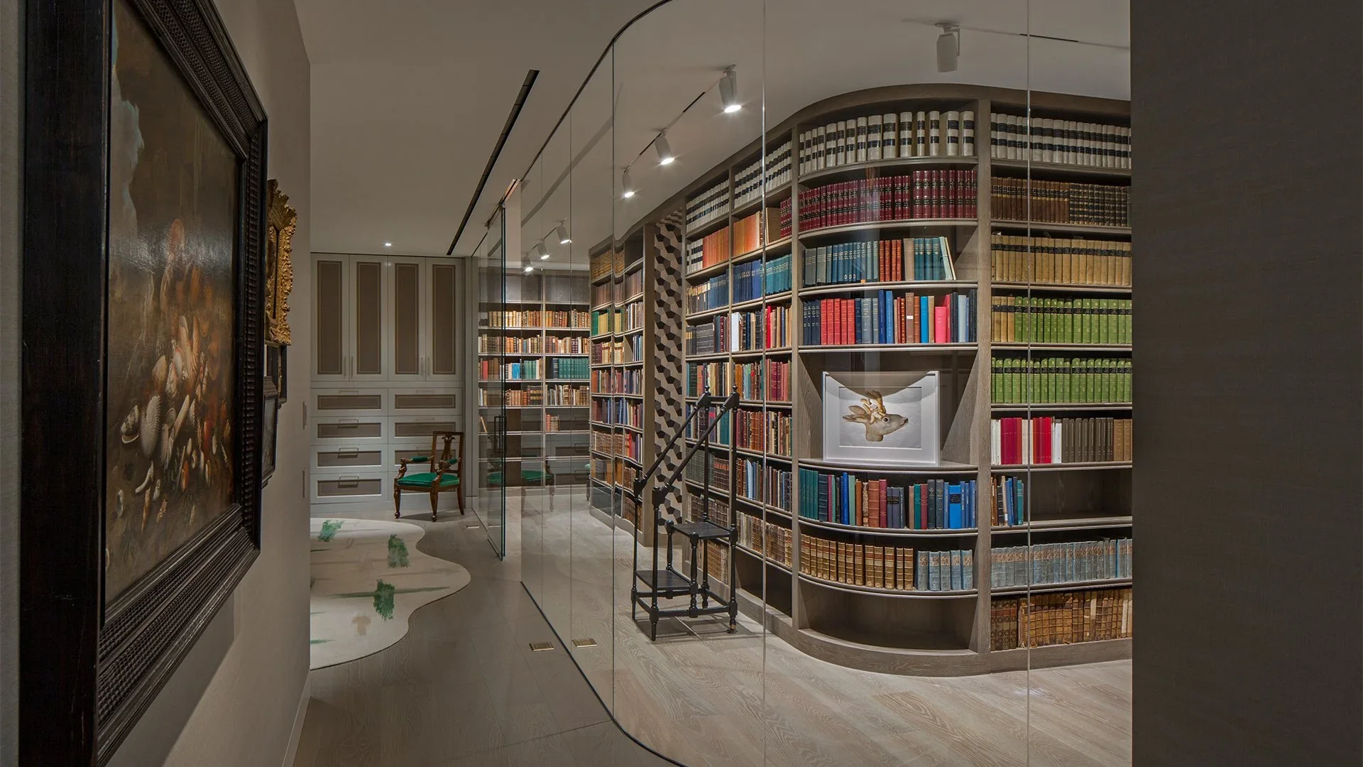 Contemporary home library with curved floor-to-ceiling bookshelves filled with colorful books, a small framed rabbit artwork, a vintage chair with green cushion, a patterned wall, and a glass wall partition.