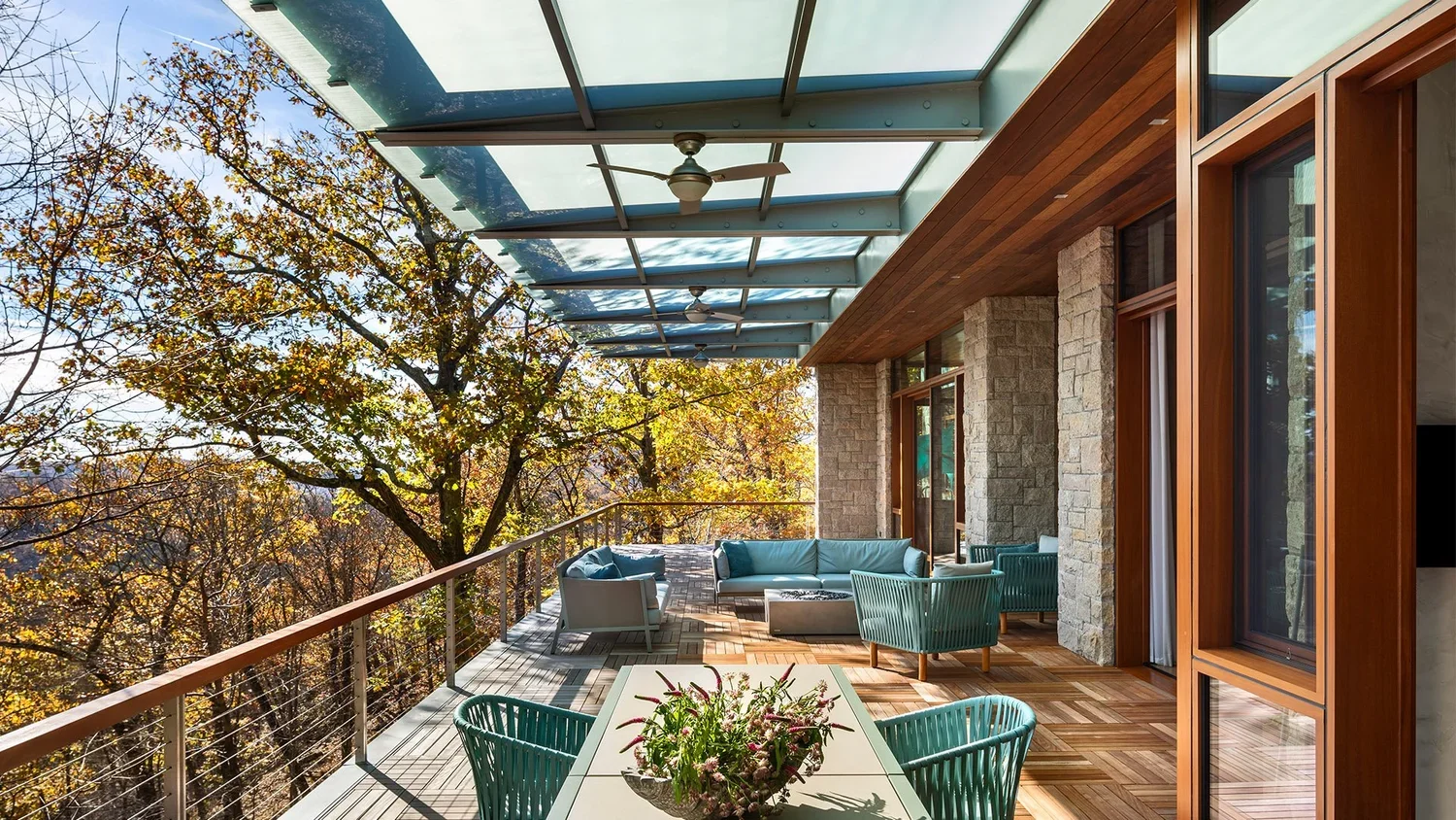 Lush outdoor balcony with wooden flooring, comfortable seating including sofas and chairs, a white dining table with a floral centerpiece, surrounded by autumn-colored trees, shaded by a glass roof with ceiling fans.