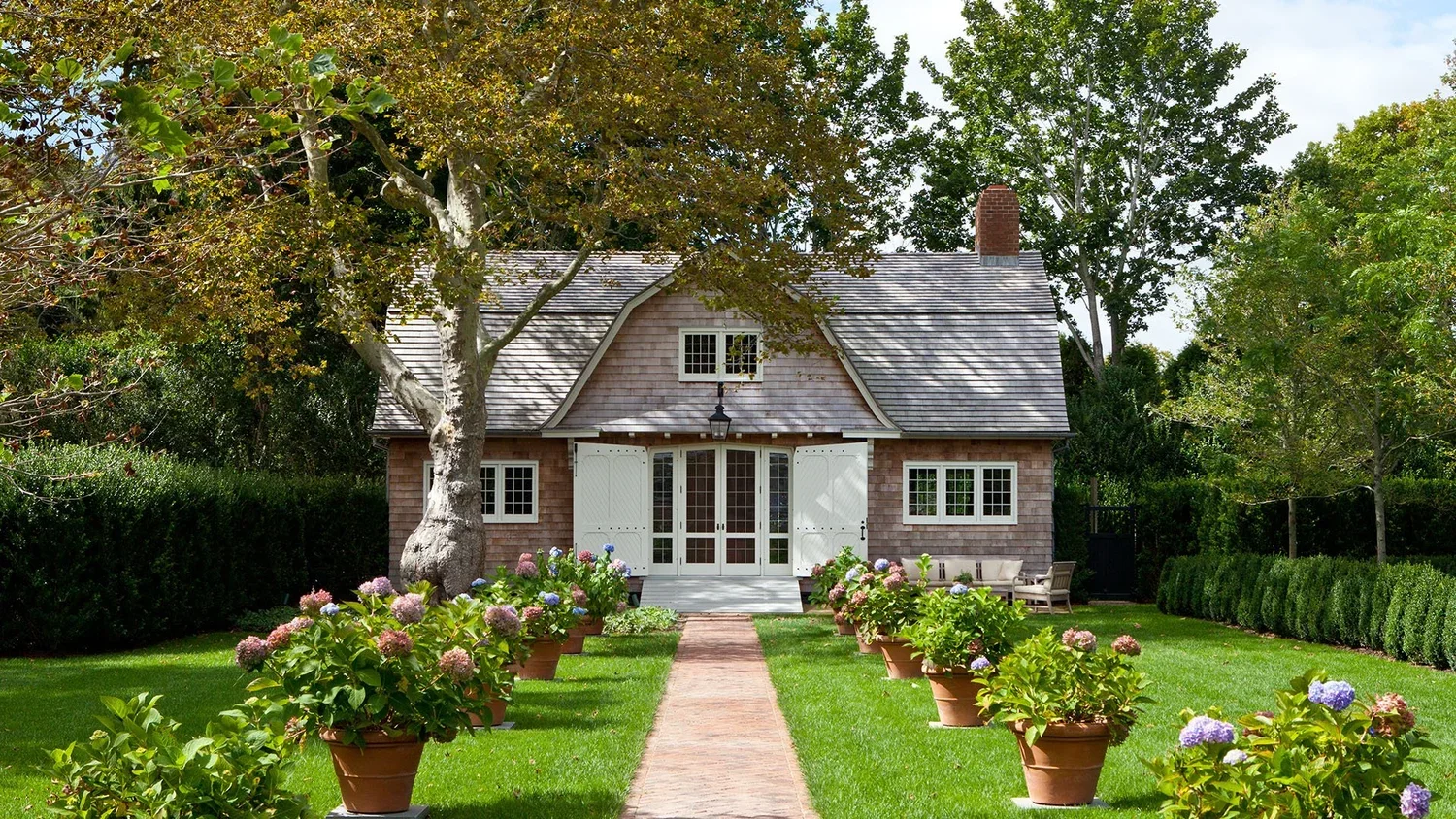A charming house with a large tree in front, surrounded by lush greenery and flowering plants, with a brick pathway leading to the front door.