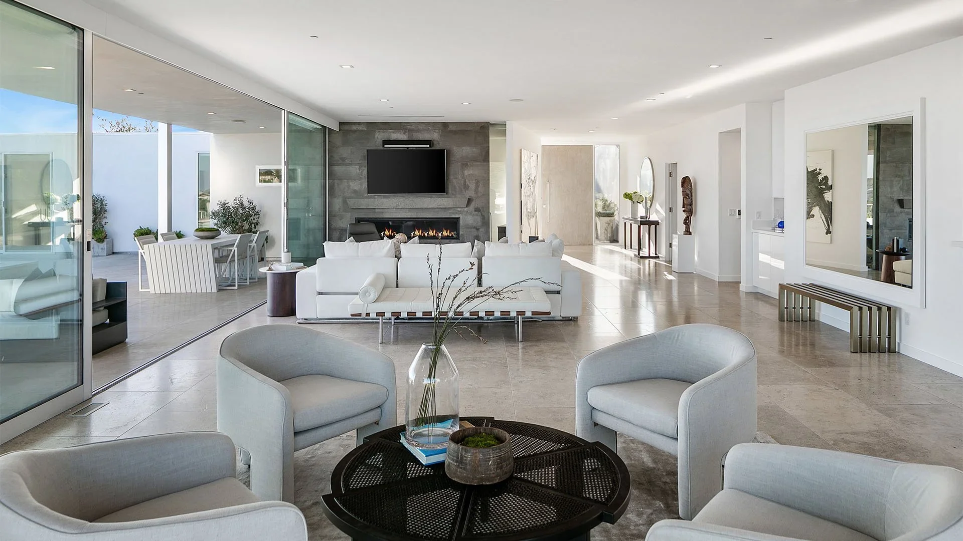 Modern living room with white sofas, chairs, a central round table with plants, and a large glass sliding door leading to an outdoor patio with dining table and potted plants.