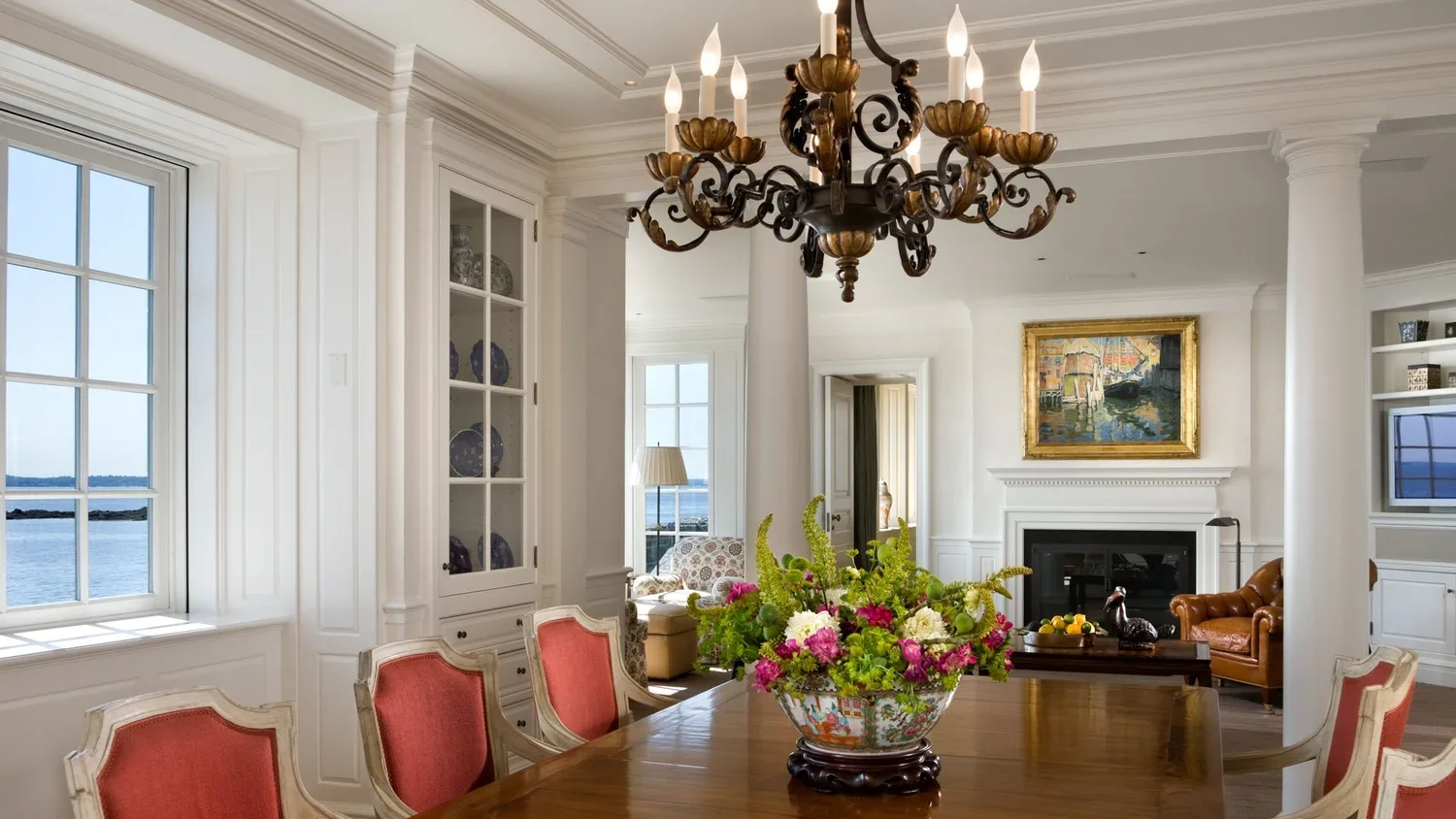 Elegant dining room with a large wooden table, a colorful floral centerpiece, and a vintage chandelier hanging from the ceiling. The room features large windows with a view of the water, white walls, and built-in display cabinets.