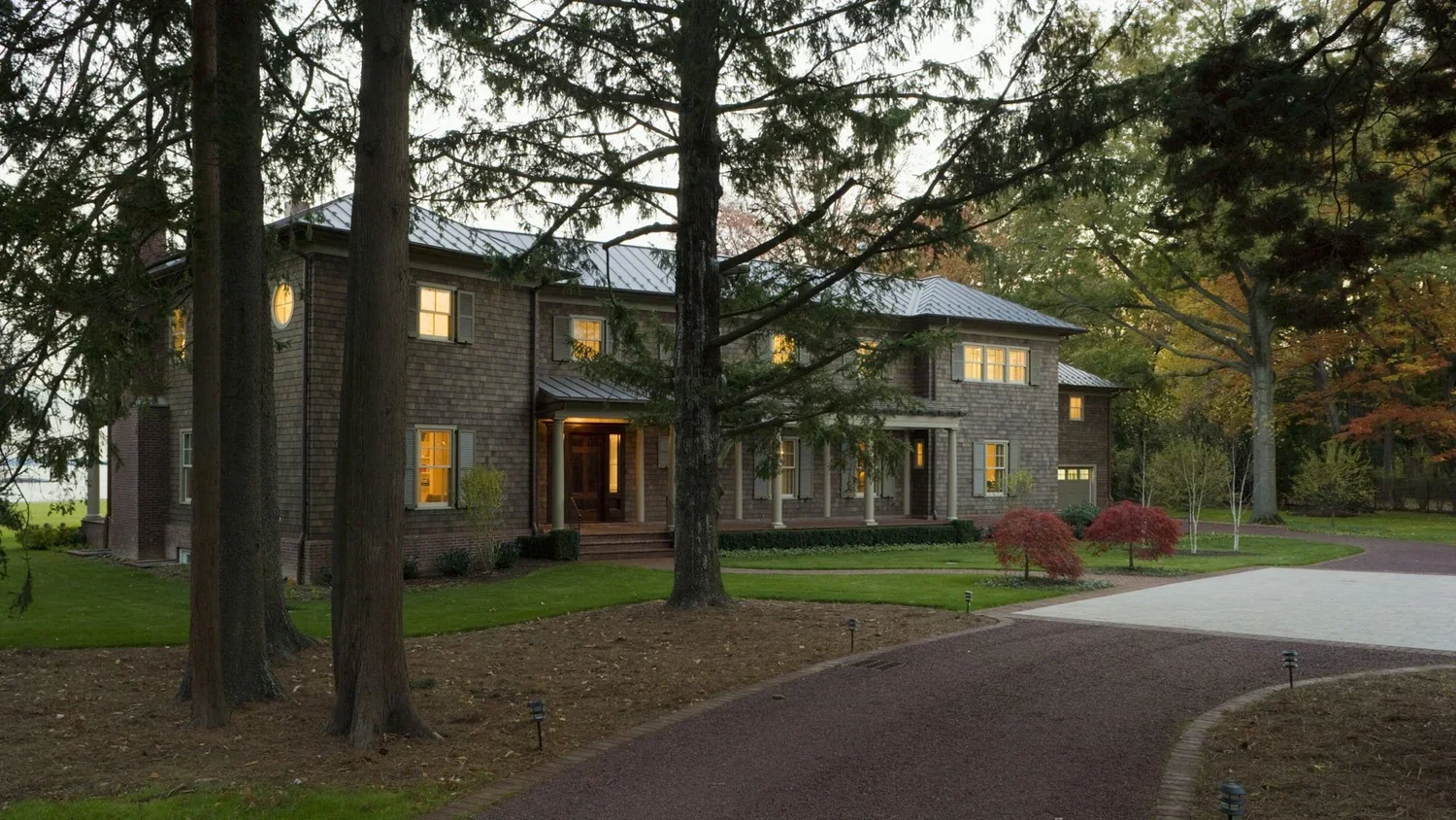Large house surrounded by trees with lights on inside, landscaped yard with walkway, grass, and small planted trees.