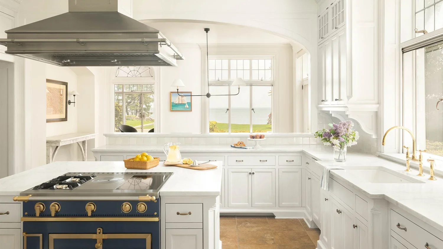 Bright white kitchen with large windows overlooking a grassy landscape and water, featuring a black and gold stove, white cabinetry, a white countertop, a vase with flowers, and a basket of lemons.