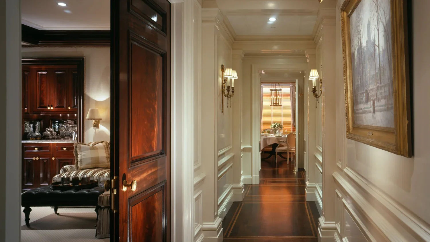 Elegant hallway with white walls, decorative moldings, wall sconces, and a large framed painting. At the end of the hall, there is a well-lit dining area with a round table, chairs, and a window with blinds.