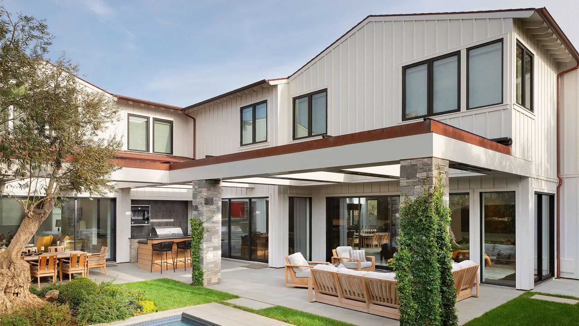 Modern multi-story house with a patio featuring outdoor seating, a barbecue grill, and a landscaped yard with grass, trees, and a swimming pool.