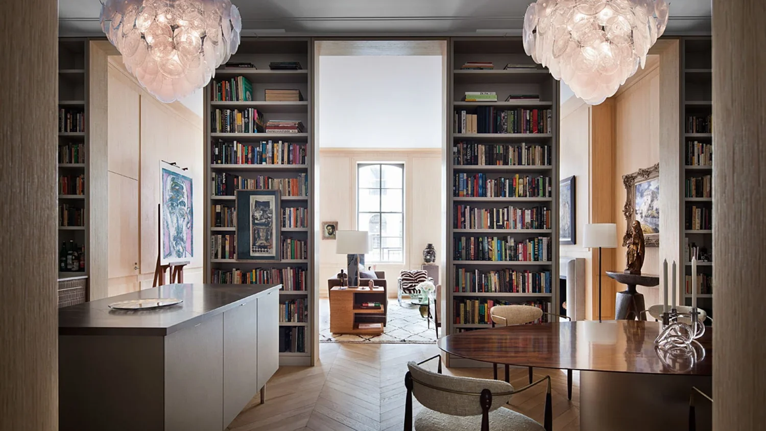 View of a stylish home library with floor-to-ceiling bookshelves, a wooden table, and modern chandeliers, leading to a bright sitting area with large window.