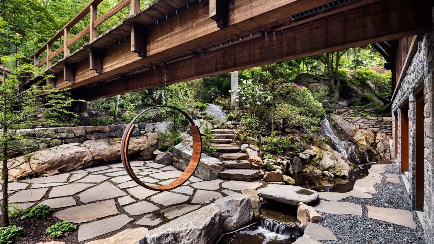 A backyard with a wooden bridge, a circular wooden swing, stone pathway, small waterfall, stone steps, and lush green trees and plants.