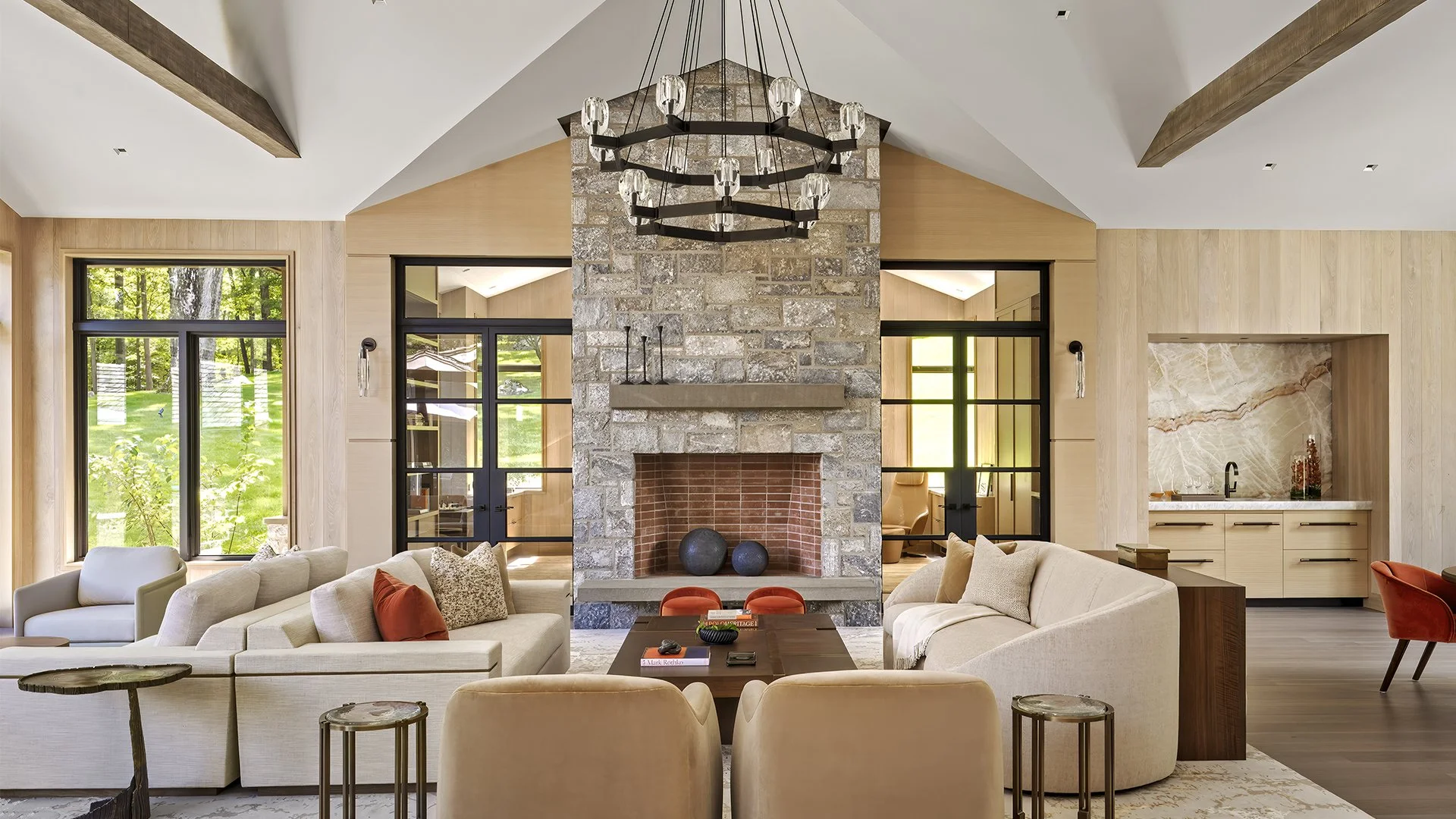 Living room with beige sofas, decorative cushions, a dark wooden coffee table, a stone fireplace with two decorative spheres, large windows showing greenery outside, a modern chandelier hanging from a vaulted ceiling, and a kitchenette with marble co