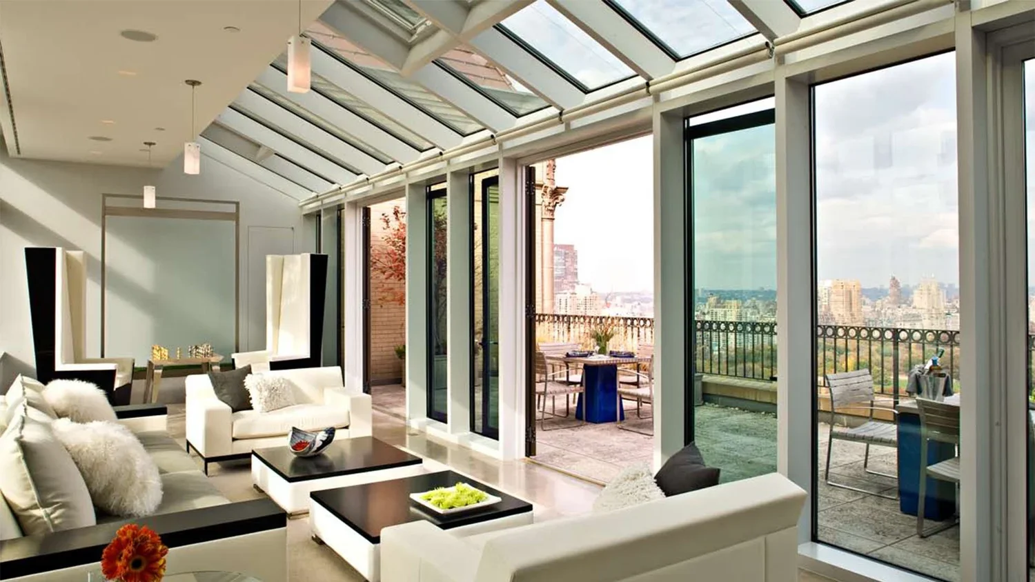 Living room with large glass windows and patio doors leading to a balcony with outdoor seating, topped with a glass ceiling allowing natural light.