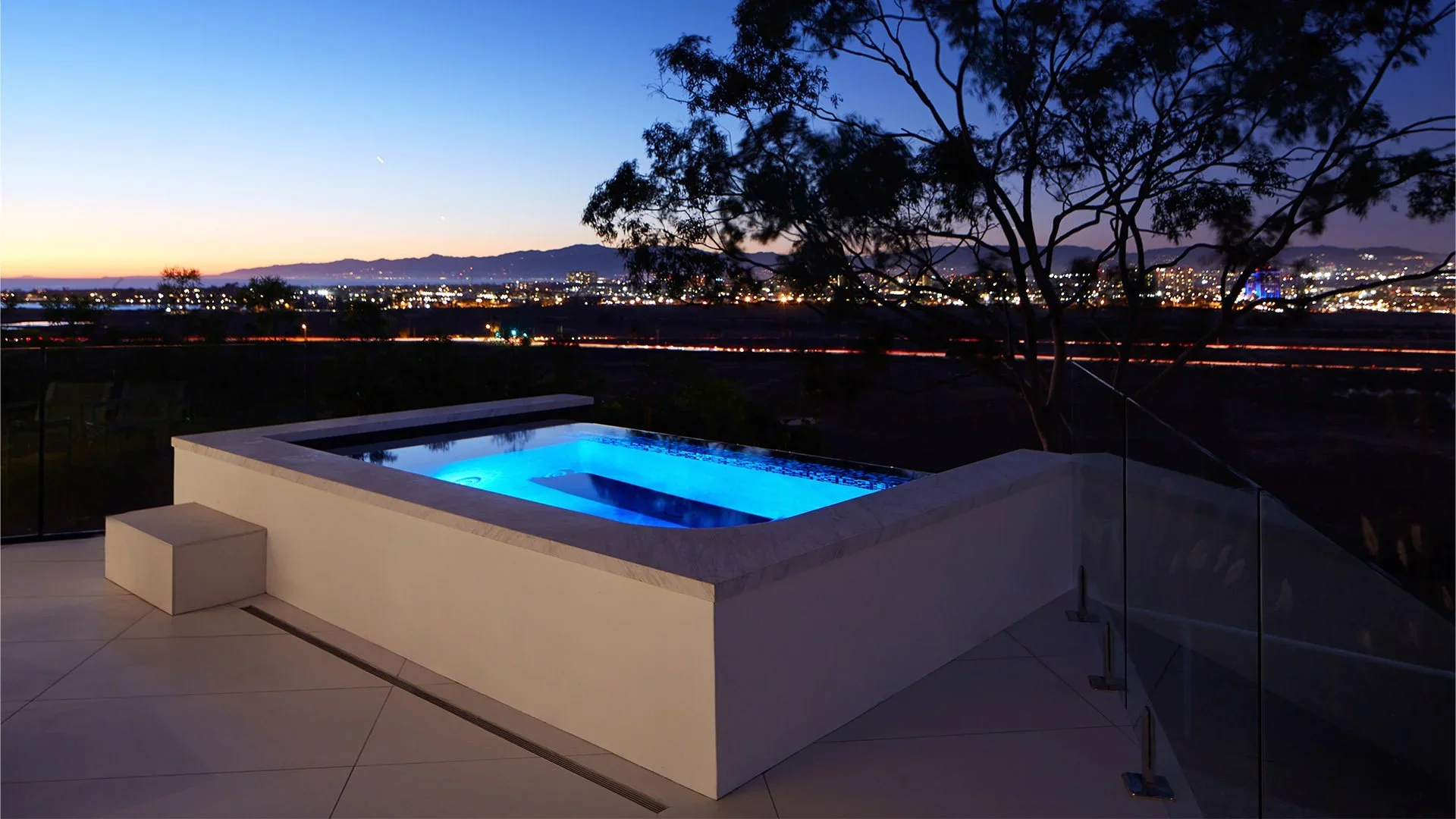 Outdoor hot tub with blue lighting on a patio, overlooking a city skyline at dusk with mountains in the distance and a clear sky.