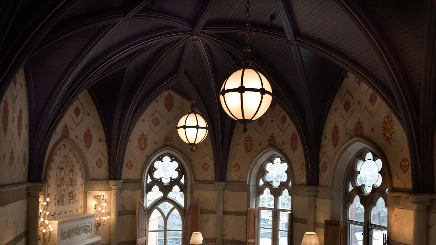 Interior of a Gothic-style room with vaulted dark wood ceiling, large stained glass windows, hanging globe chandeliers, and decorated walls with floral patterns.