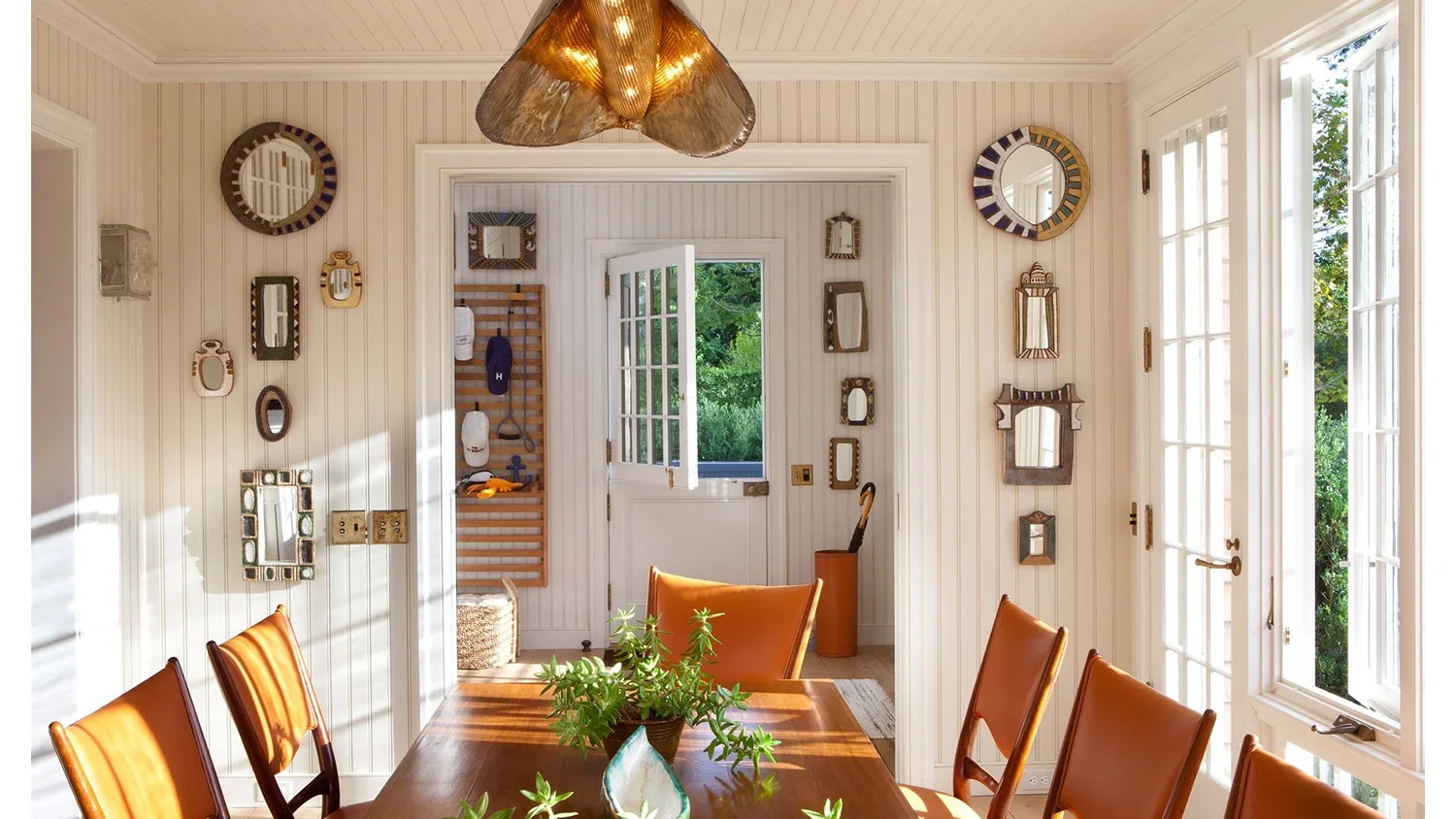 Sunlit dining room with white paneled walls, decorated with circular and rectangular mirrors, featuring a wooden dining table, tan leather chairs, a green plant centerpiece, and an open door leading outside with visible trees.