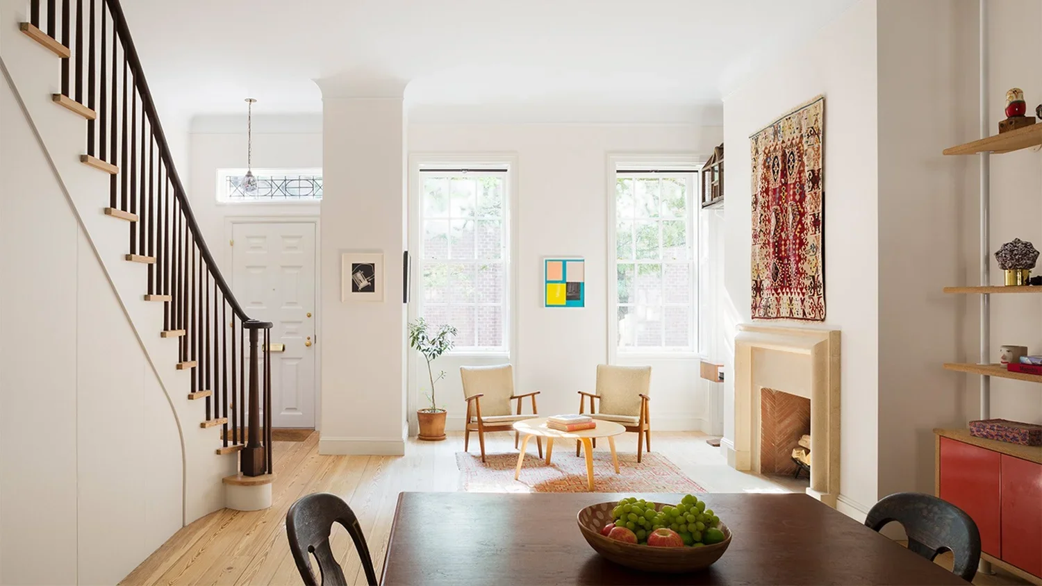 Living room with two armchairs, fireplace, colorful artwork, and windows; wooden staircase and dining table with fruit bowl in foreground.