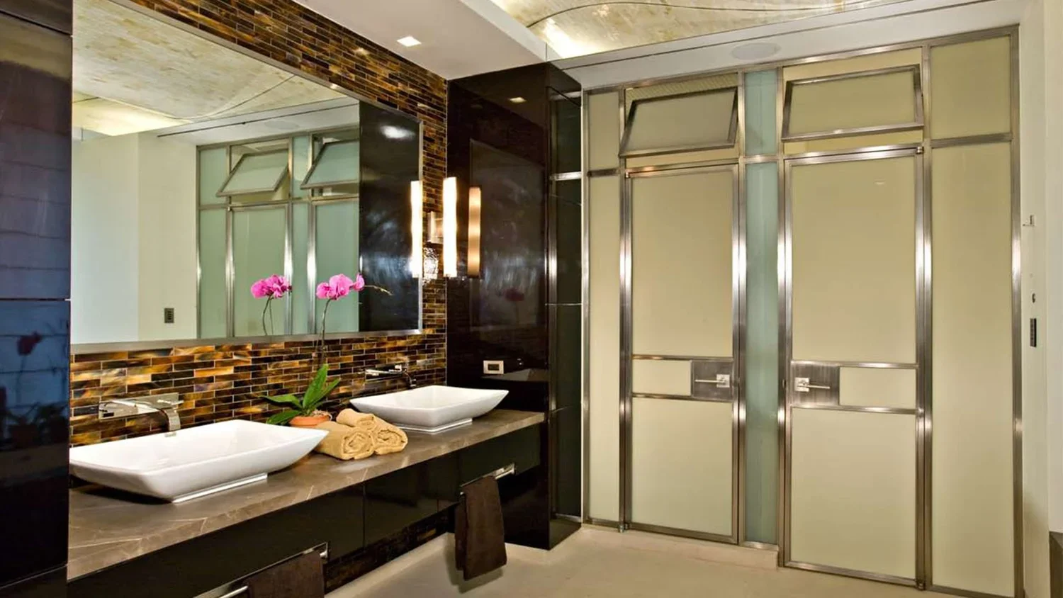 Modern hotel bathroom featuring dual vessel sinks, a large mirror, pink orchids, a colorful tile backsplash, and a frosted sliding door for the shower.