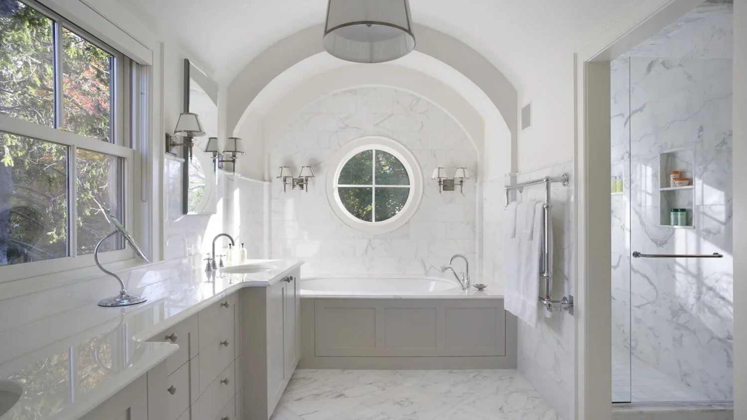 Bright, elegant bathroom with white marble walls and floors, large windows, a bathtub, and a walk-in shower.