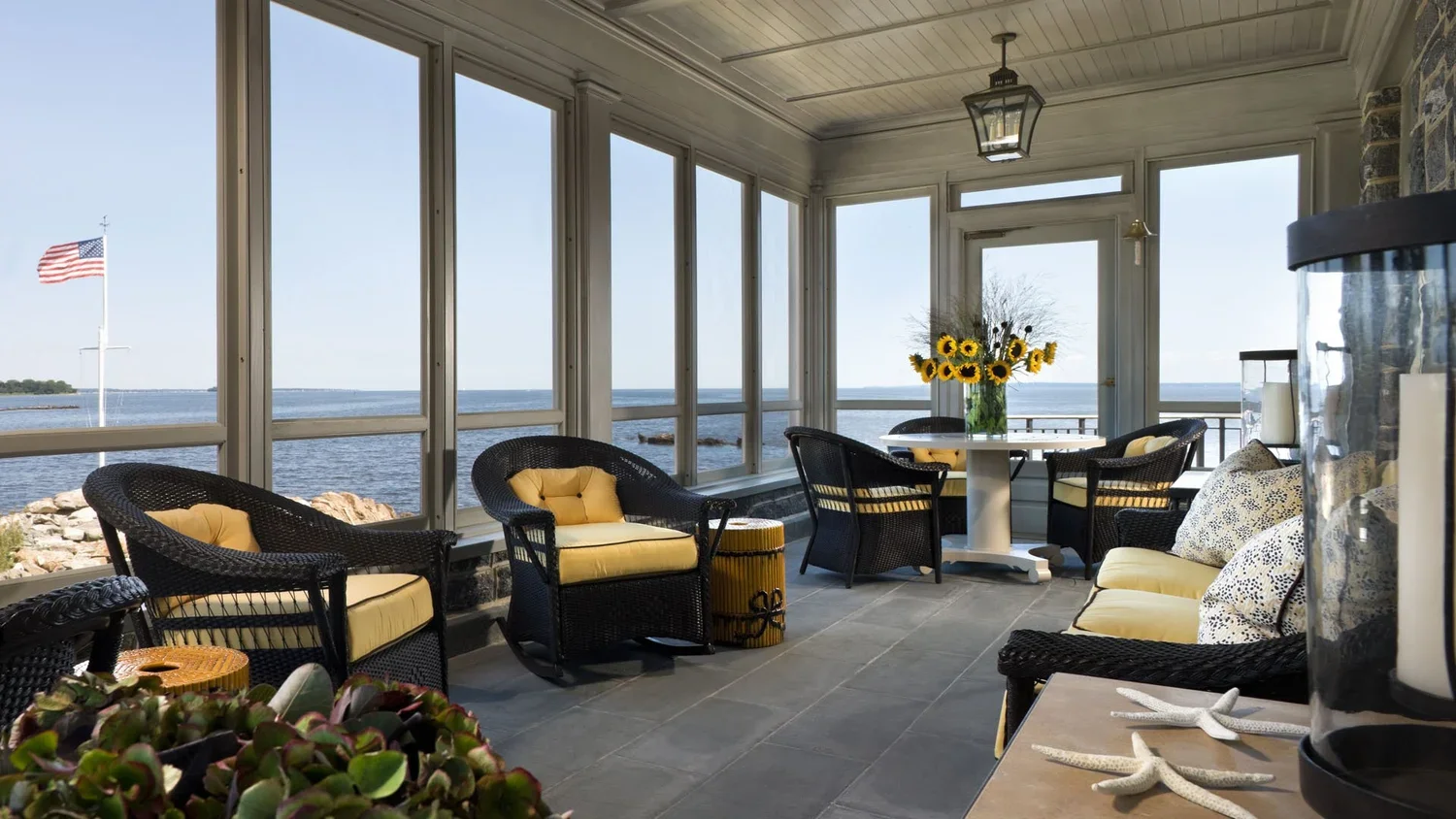 Sunroom with black wicker furniture, yellow cushions, and a vase of sunflowers overlooking a body of water with an American flag.