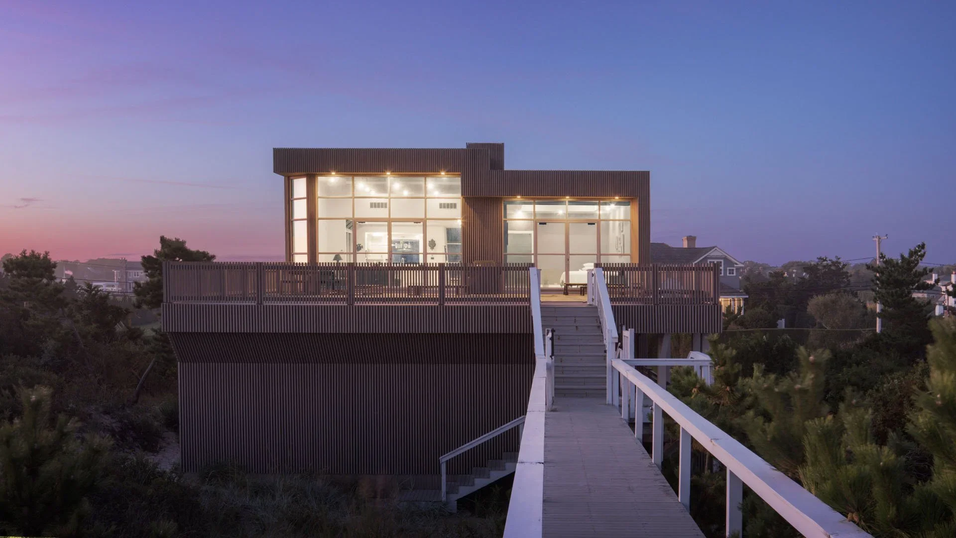 Modern house with large glass windows, wooden exterior, and a balcony, connected by a wooden ramp and stairs, set against a twilight sky with trees and neighboring houses in the background.