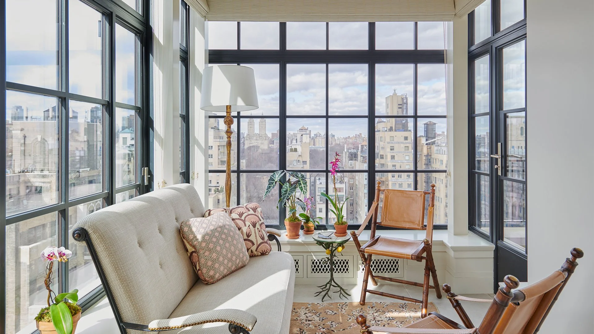 Sunlit enclosed balcony with large windows, beige sofa, potted plants, and wooden and leather chairs.